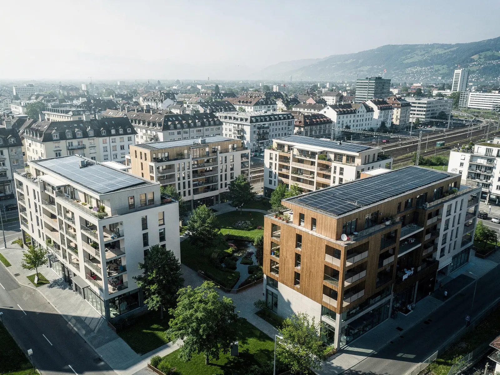Luftaufnahme eines Schweizer Areals mit sechs Wohn- und Geschäftsgebäuden um einen begrünten Innenhof, Photovoltaik auf allen Dächern