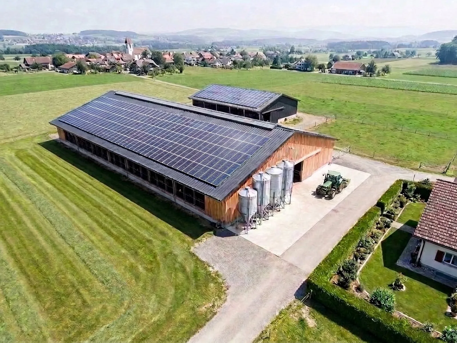 Luftaufnahme eines Schweizer Landwirtschaftsbetriebs mit zwei Ställen, vollflächiger Photovoltaik auf beiden Dächern, Futtersilos und Dorf im Hintergrund