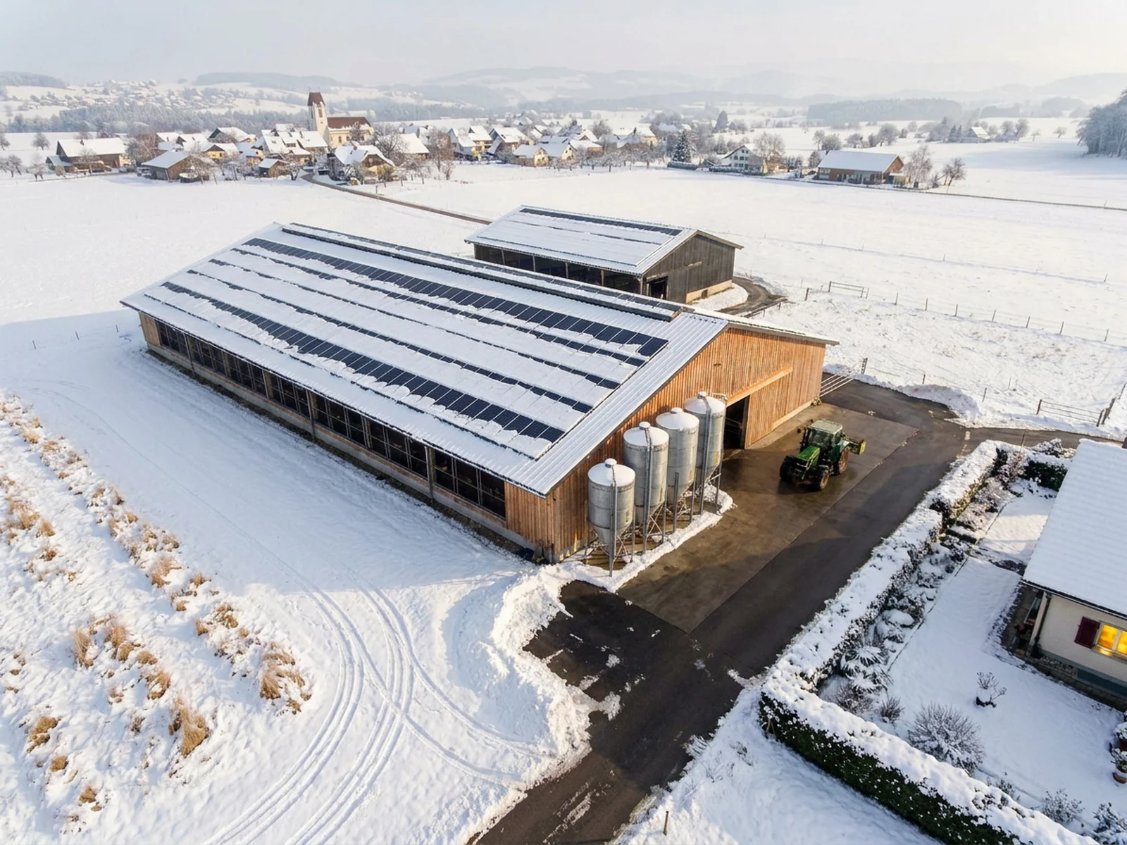 Luftaufnahme eines Schweizer Landwirtschaftsbetriebs mit zwei Ställen, vollflächiger Photovoltaik auf beiden Dächern, Futtersilos und Dorf im Hintergrund, im Winter
